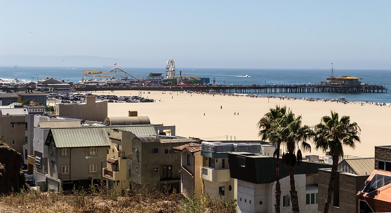 A wide sandy beach with a long pier and a Ferris wheel in the distance, and rooftops and palm trees in the foreground.