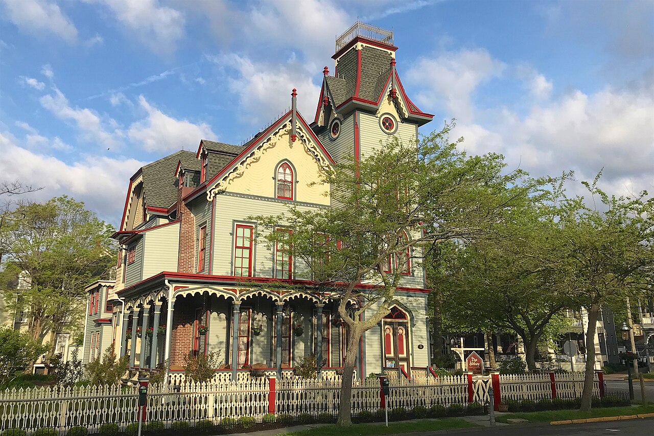 A pale green Victorian house with a tall tower, red trim and a wraparound porch behind a white picket fence.