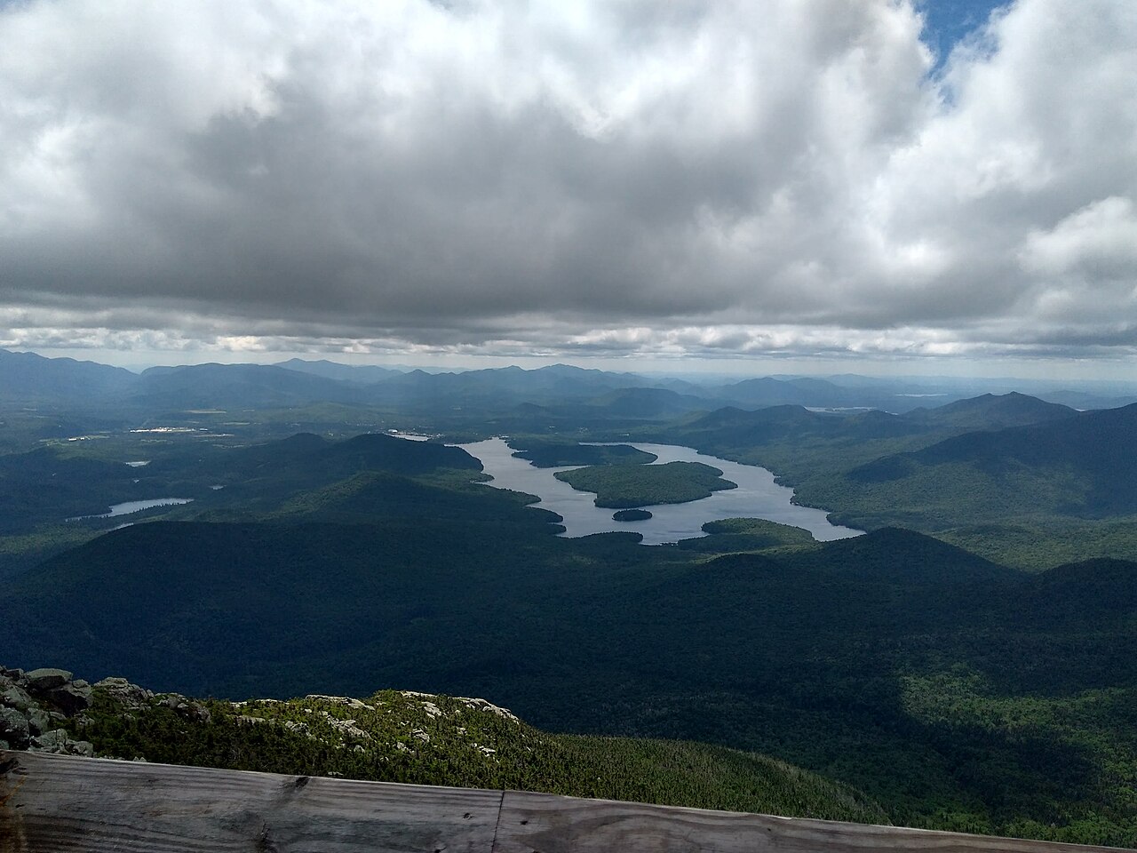 A long lake with wooded islands lying among forested hills under heavy grey clouds, seen from a mountain top.
