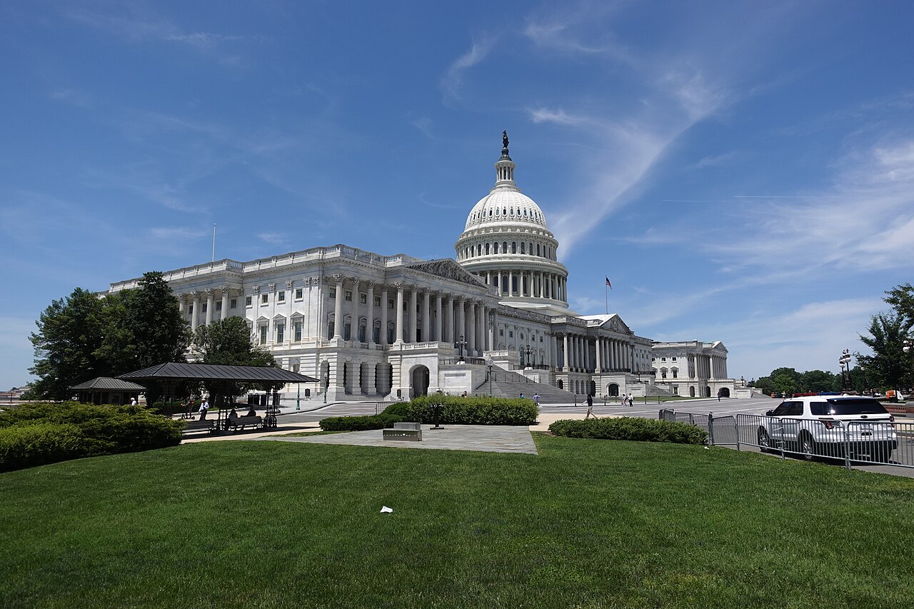 A white domed building with columned wings seen across a green lawn under a blue sky.