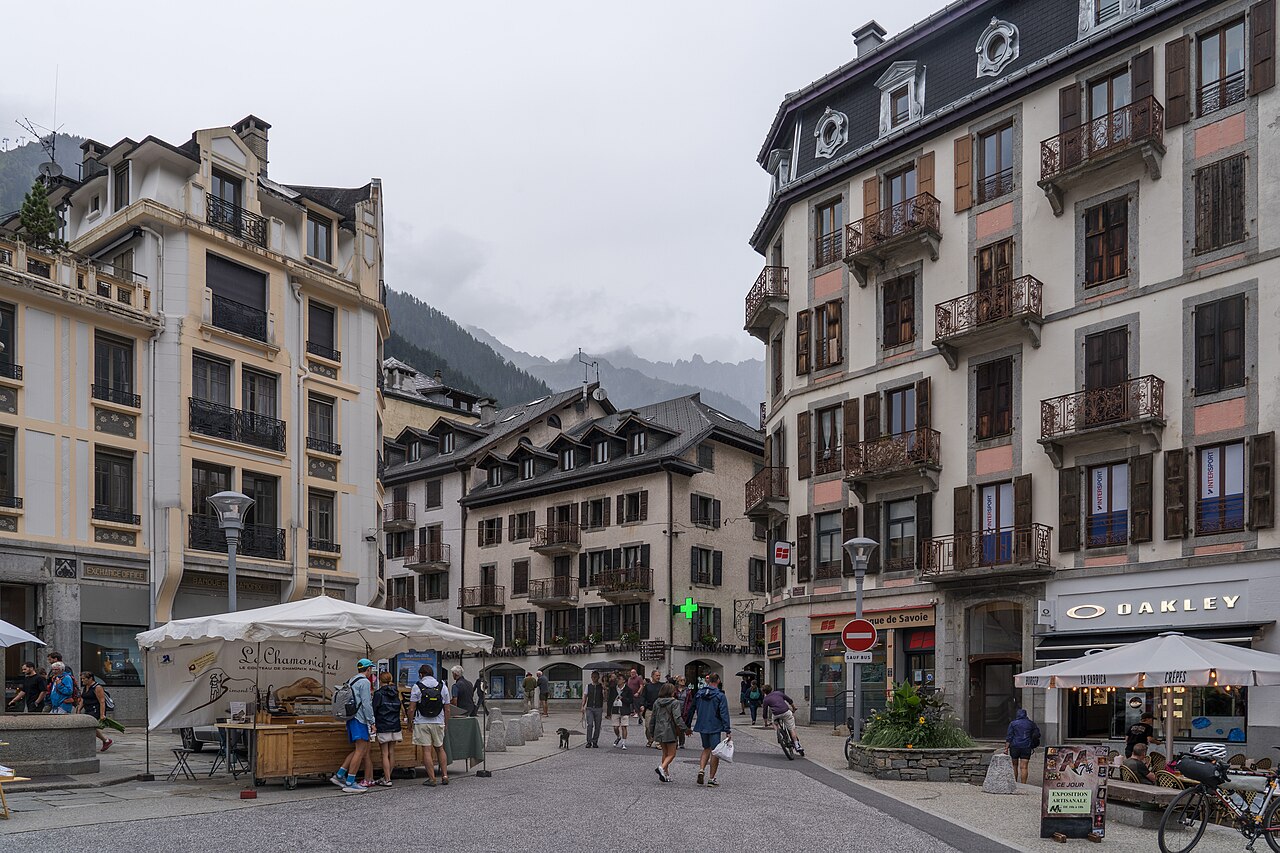 A paved town square lined with tall shuttered buildings with balconies, with a market stall and a few distant walkers.