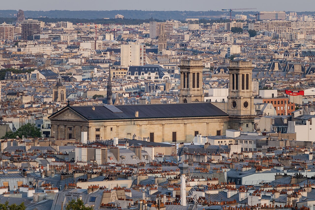 A dense spread of grey zinc roofs and chimneys with a twin-towered church rising in the middle.