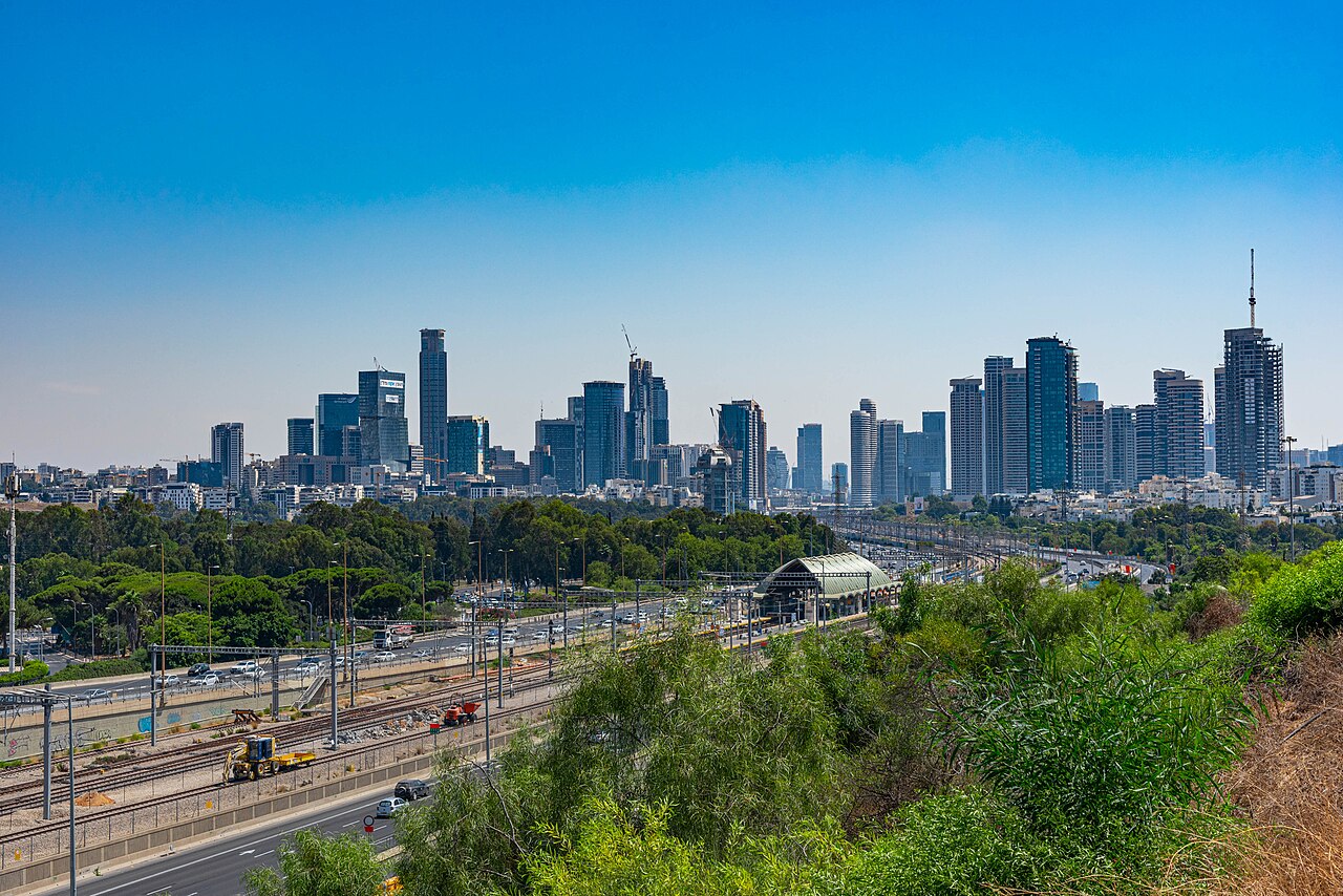 A line of glass towers on the horizon above green parkland and a railway, under a clear blue sky.