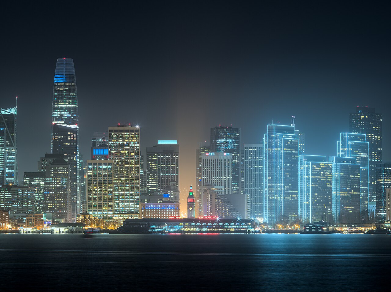 Lit skyscrapers glowing blue through fog above dark water at night.
