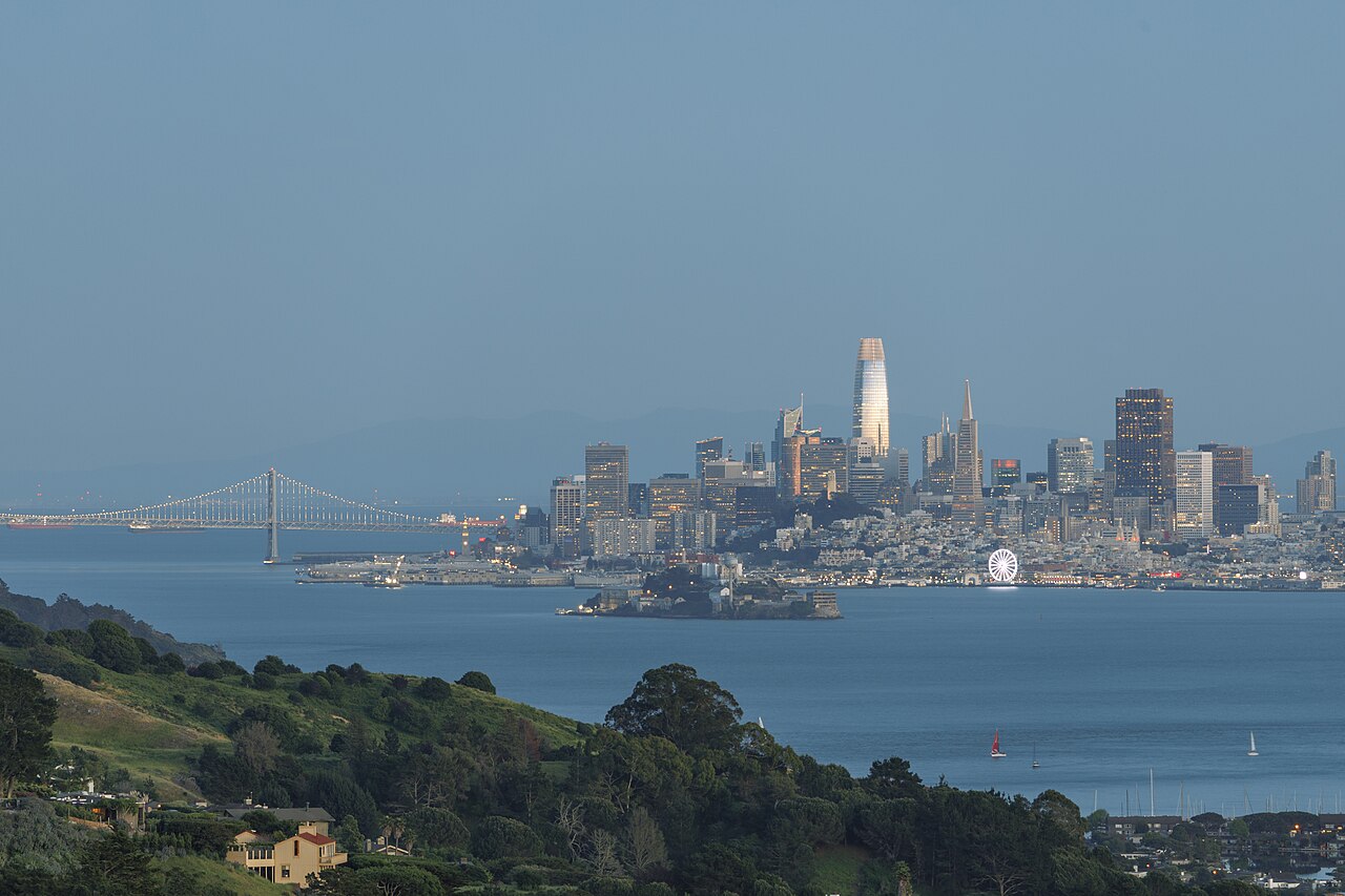 A distant city skyline across blue water, with a suspension bridge on the left and green hills in the foreground.