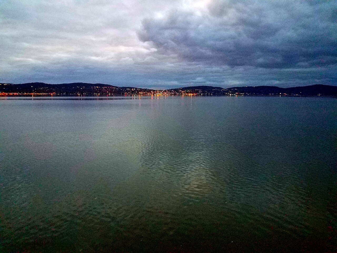 A wide stretch of calm river under heavy clouds, with town lights along the far wooded shore.