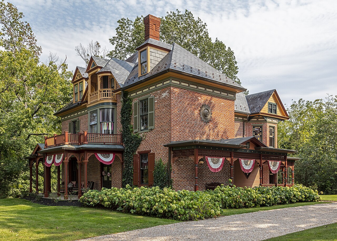 A red brick Victorian house with a tall chimney, ornate porches and flag bunting, set in a green garden.