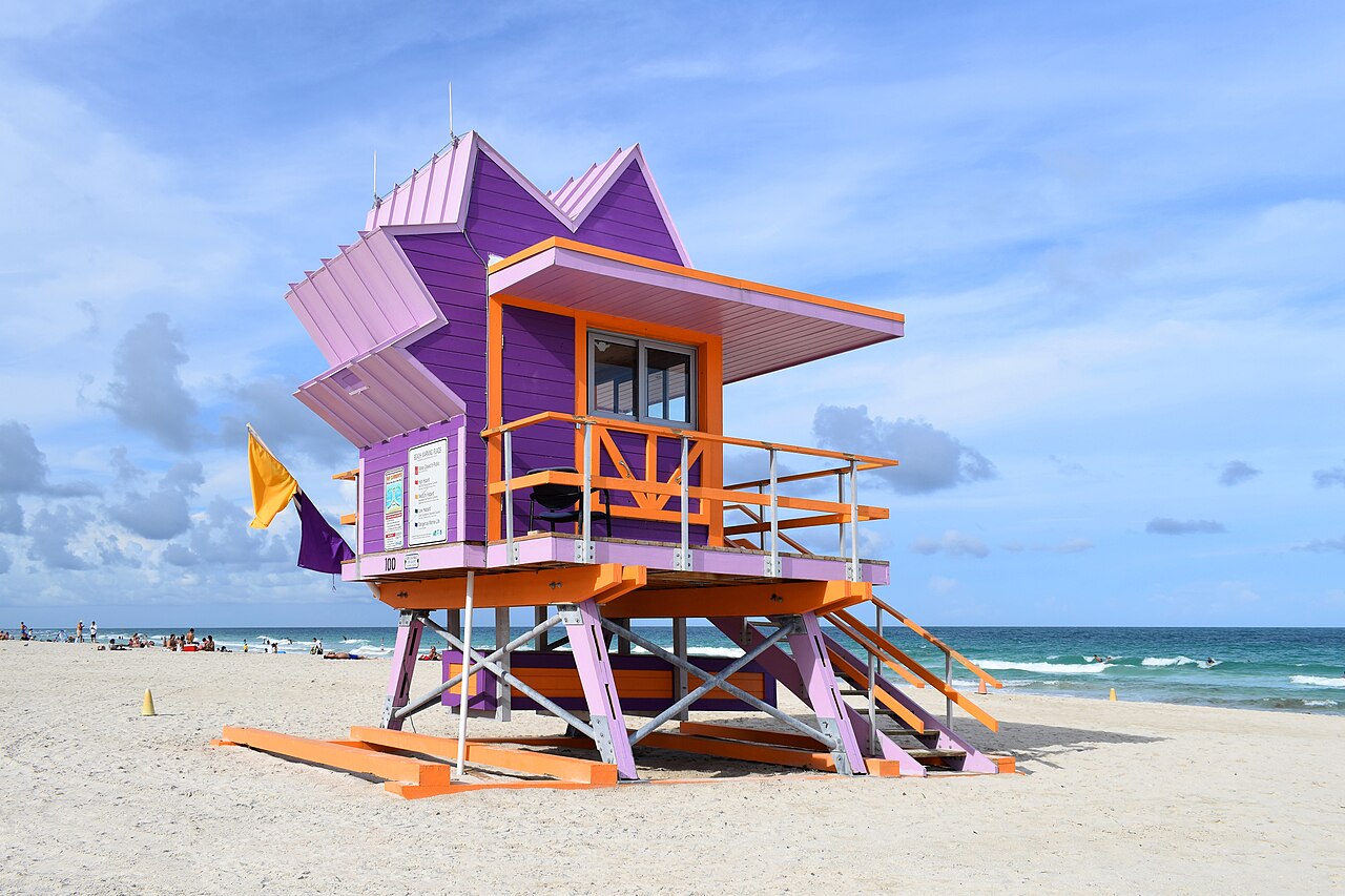 A purple and orange lifeguard tower with a jagged roofline standing on pale sand beside a turquoise sea.