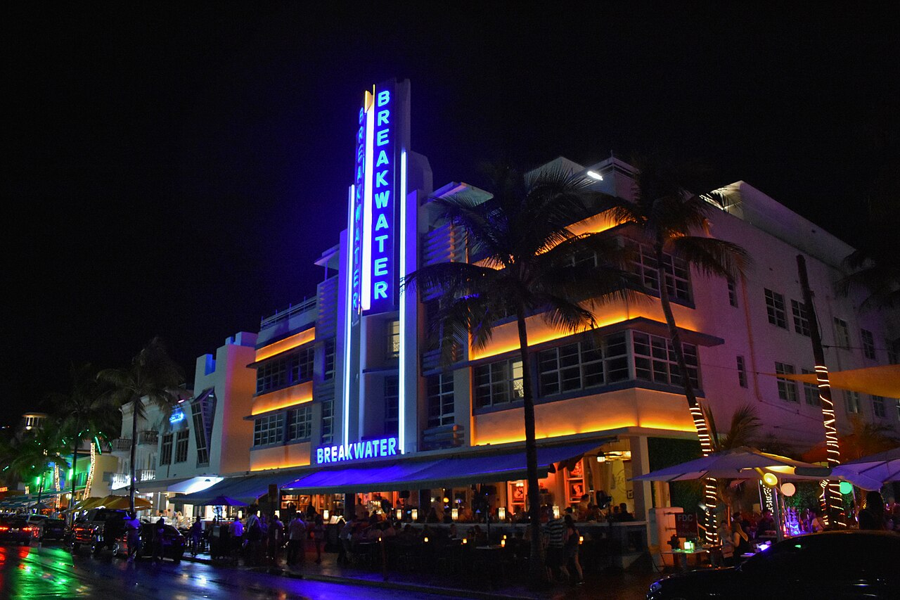A white Art Deco hotel at night with a tall central tower outlined in blue neon and orange light along its balconies.