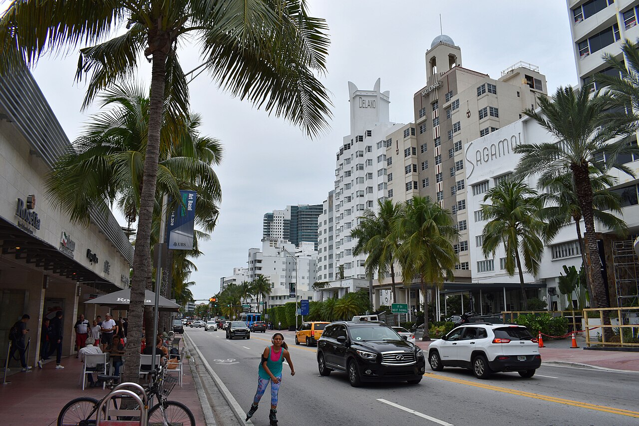 A palm-lined avenue with a row of tall pale hotel towers, cars passing and a cyclist at the kerb.