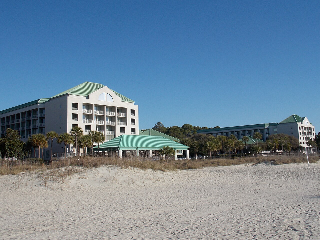 A white multi-storey resort hotel with green roofs set behind dunes and a wide empty beach.