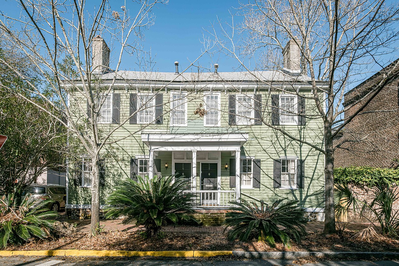 A pale green two-storey wooden house with a small columned porch, seen through bare trees and palmetto shrubs.