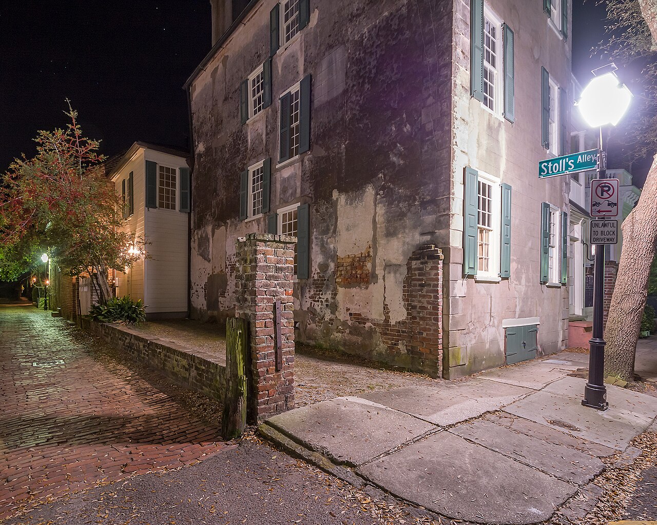 A narrow lane at night between old stuccoed houses with shutters, lit by a single bright street lamp.