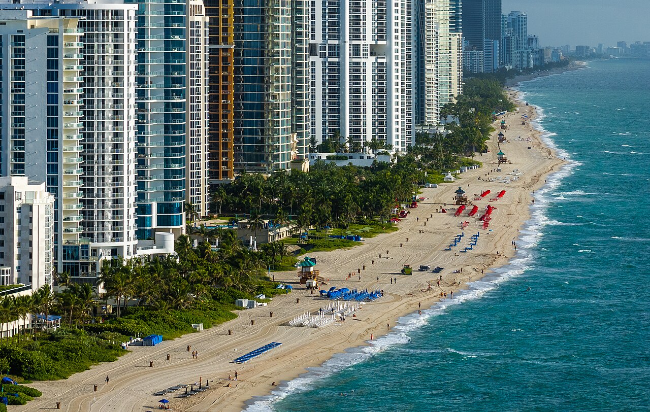 A line of glass high-rise towers beside a long sandy beach with rows of sunbeds and green-blue surf.