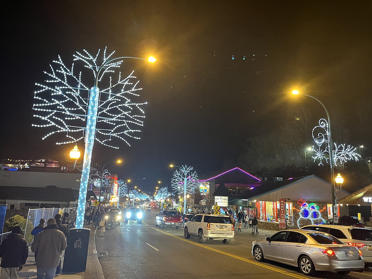 A busy street at night with trees wrapped in white lights, shopfronts, cars and people on the pavement.