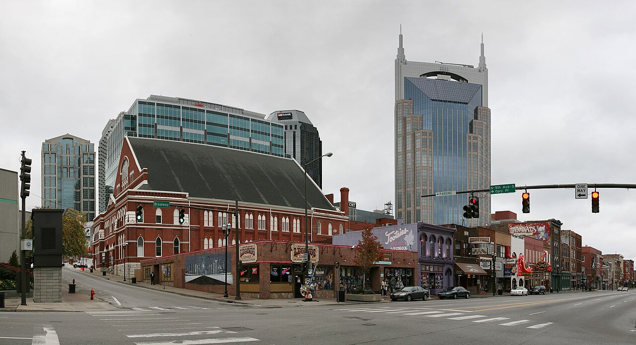 A street corner of red brick buildings with a tall glass tower topped by twin spires behind them.
