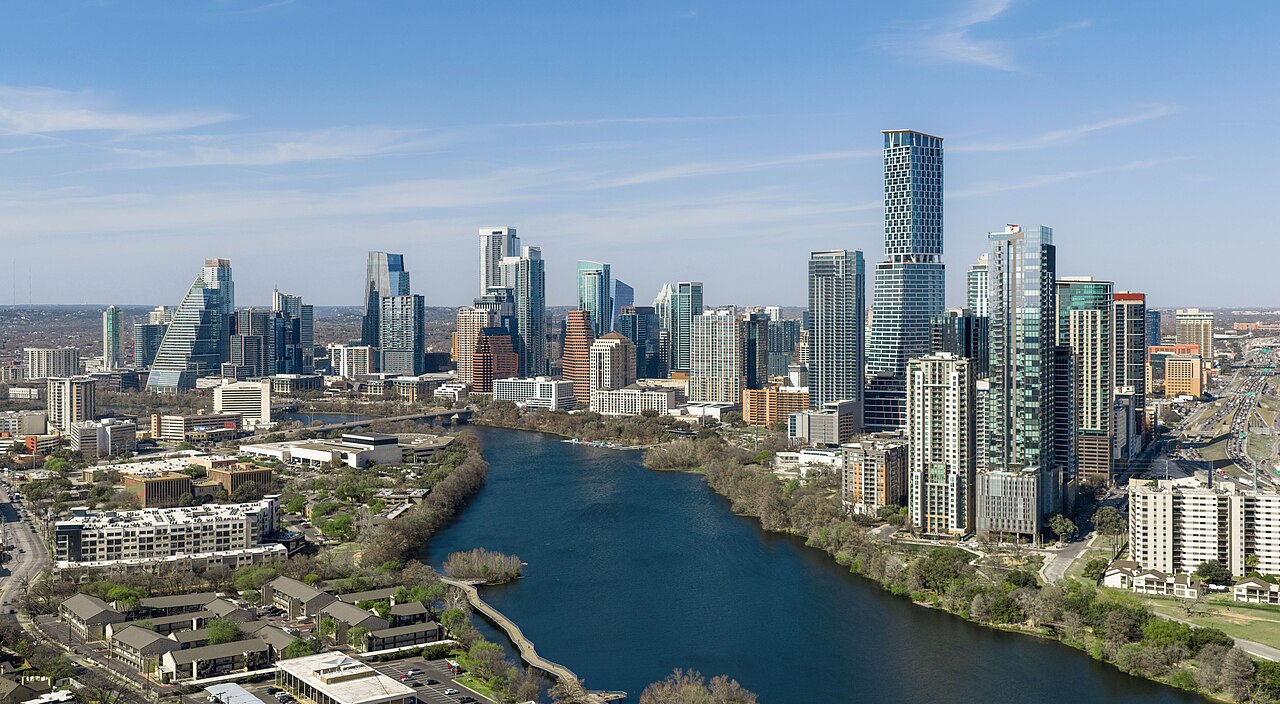 Glass high-rise towers rising behind a tree-lined river that bends through the foreground.