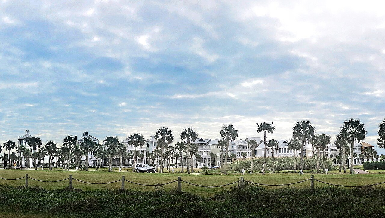 A row of large pale houses on stilts among palm trees, seen across a grassy field under a cloudy sky.