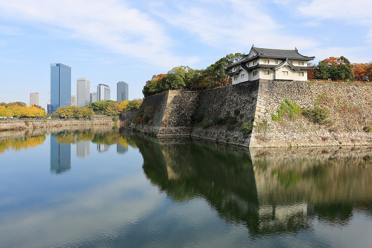 A white castle turret on a high stone wall reflected in still water, with modern office towers in the distance.