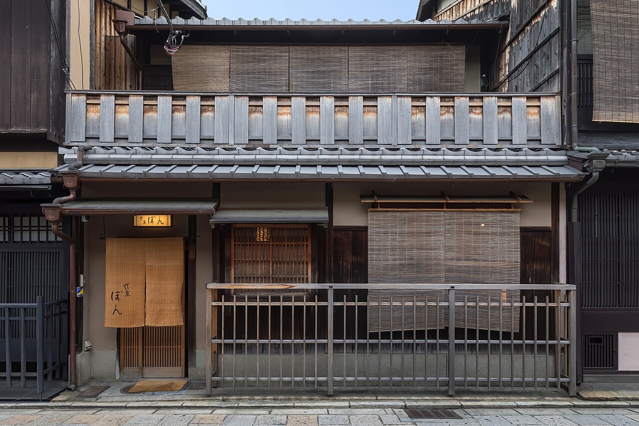 A two-storey dark timber house with reed blinds, a wooden balcony rail and a small yellow lamp over the door.