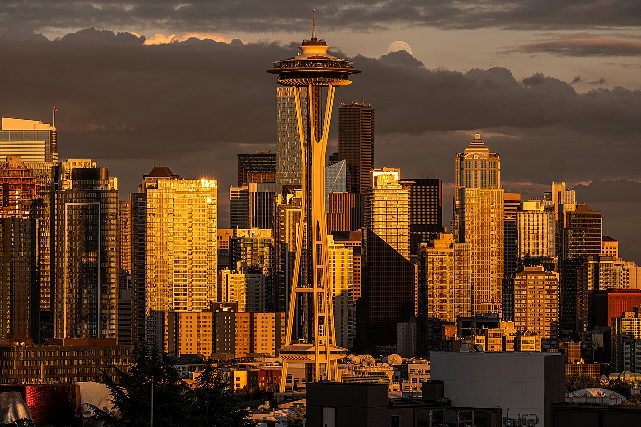 A tall observation tower in front of downtown high-rises glowing gold in low evening light.