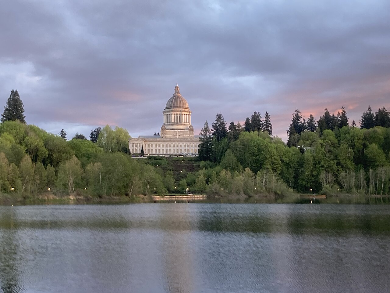 A domed stone building rising above a wooded shore, with a calm lake in front and pink clouds overhead.