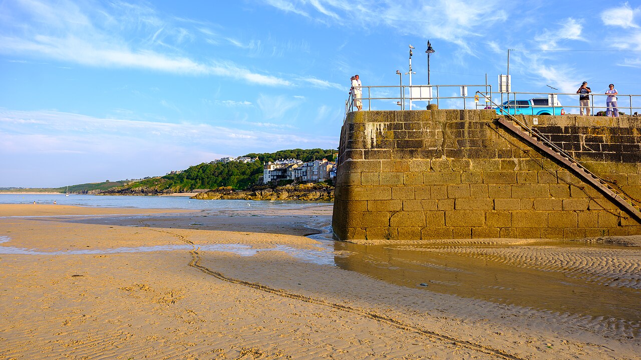 A stone pier rising from wet golden sand, with a green headland and houses across the bay.