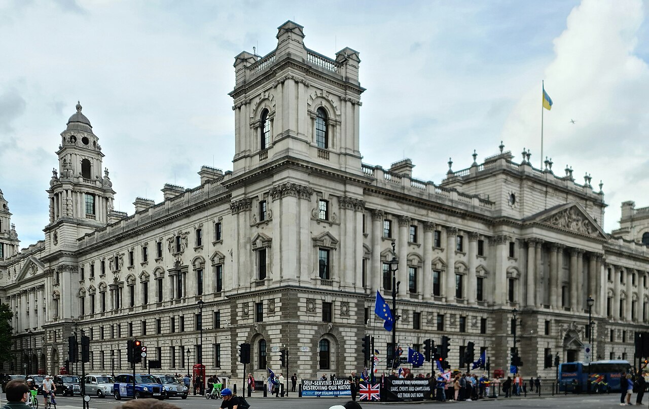 A large pale stone government building with corner towers and rows of arched windows, seen from across the street.