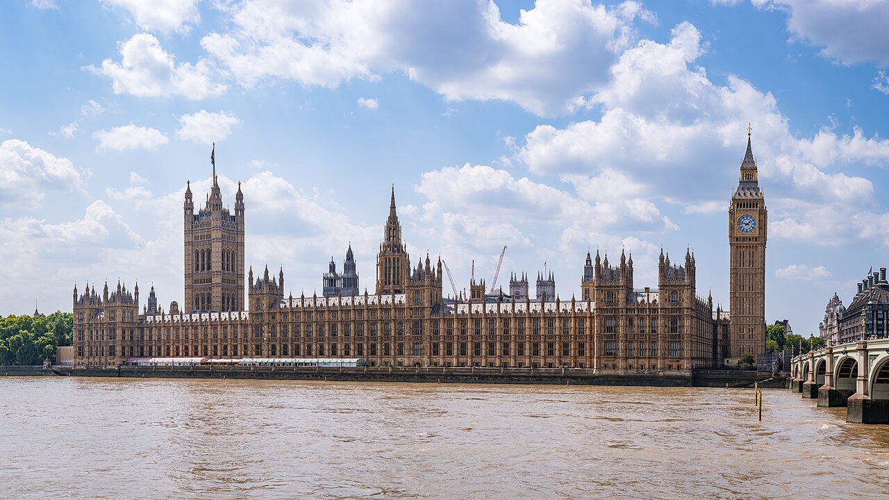 A long Gothic riverside building with a clock tower at one end, under blue sky and white clouds.