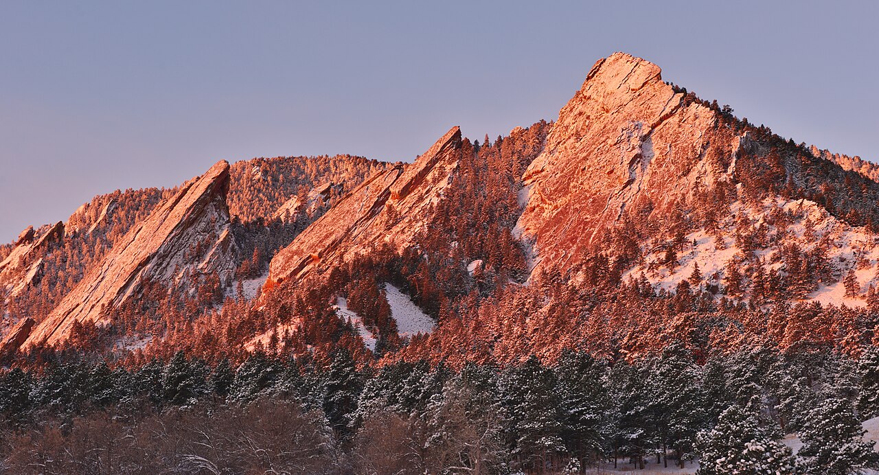 Sunlight illuminates towering sandstone rock formations rising above a snow-covered pine forest.