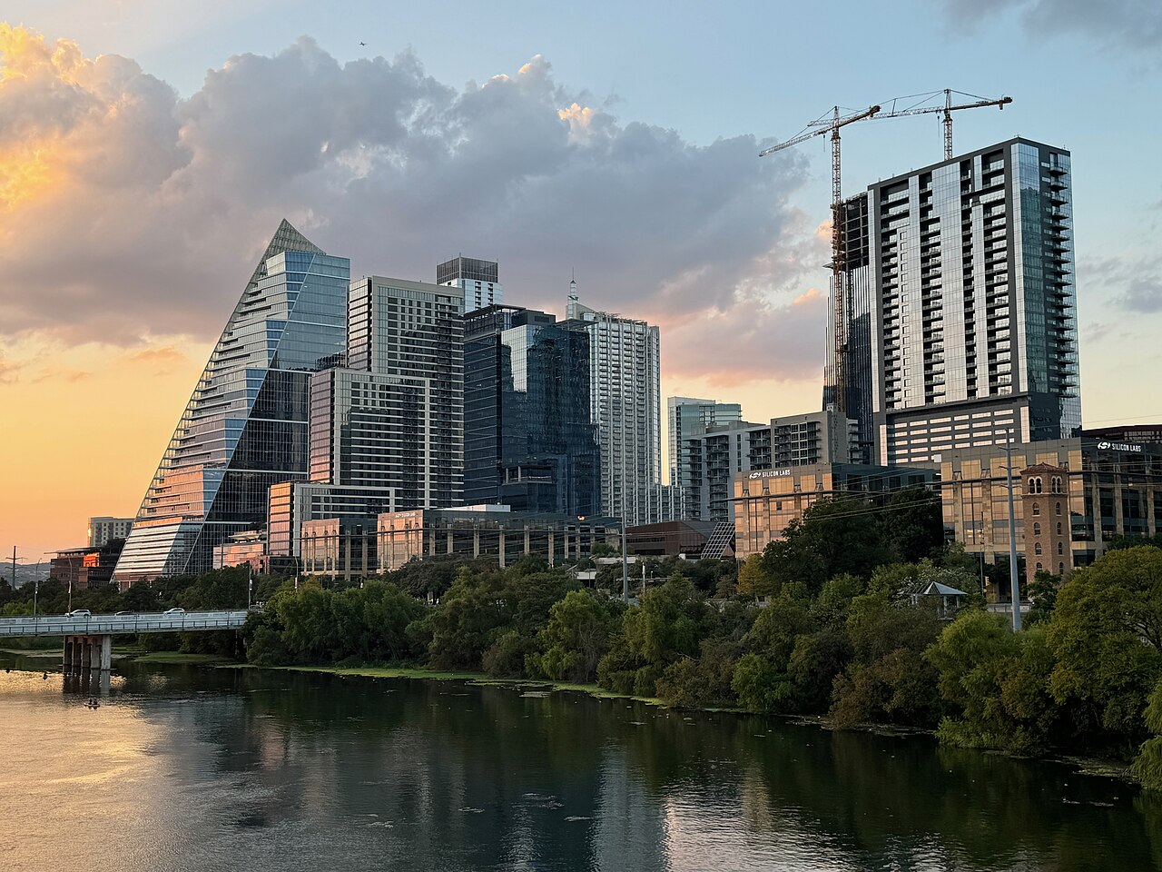 Glass towers, one with a sloping sail-shaped side, rising behind a tree-lined lake at dusk.