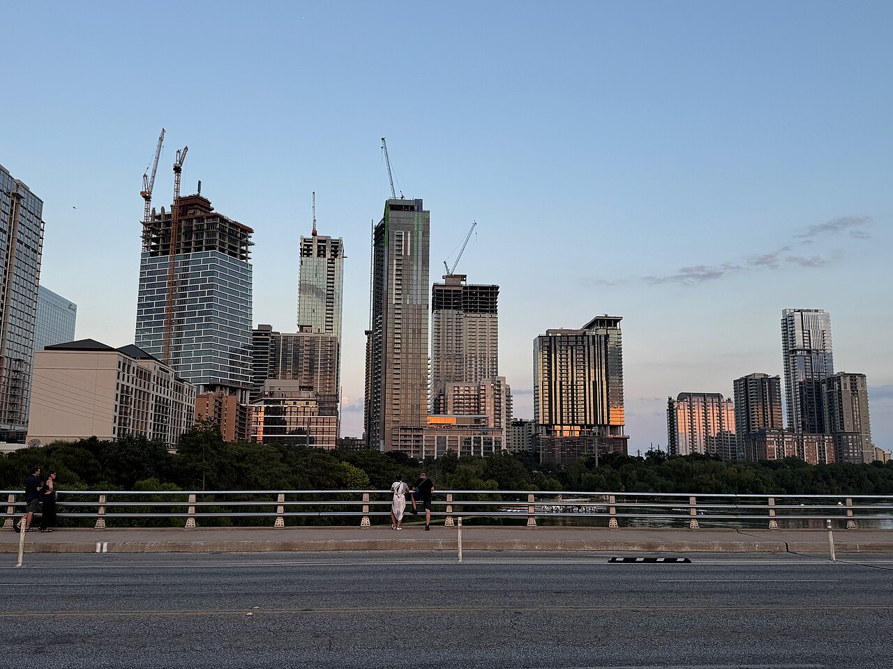 A line of slim residential towers, some with cranes on top, seen beyond a bridge railing at dusk.