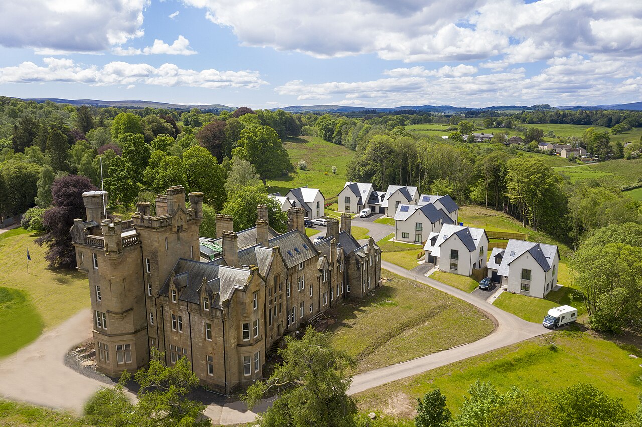 An aerial view showing a stone castle alongside new residential houses surrounded by green trees.