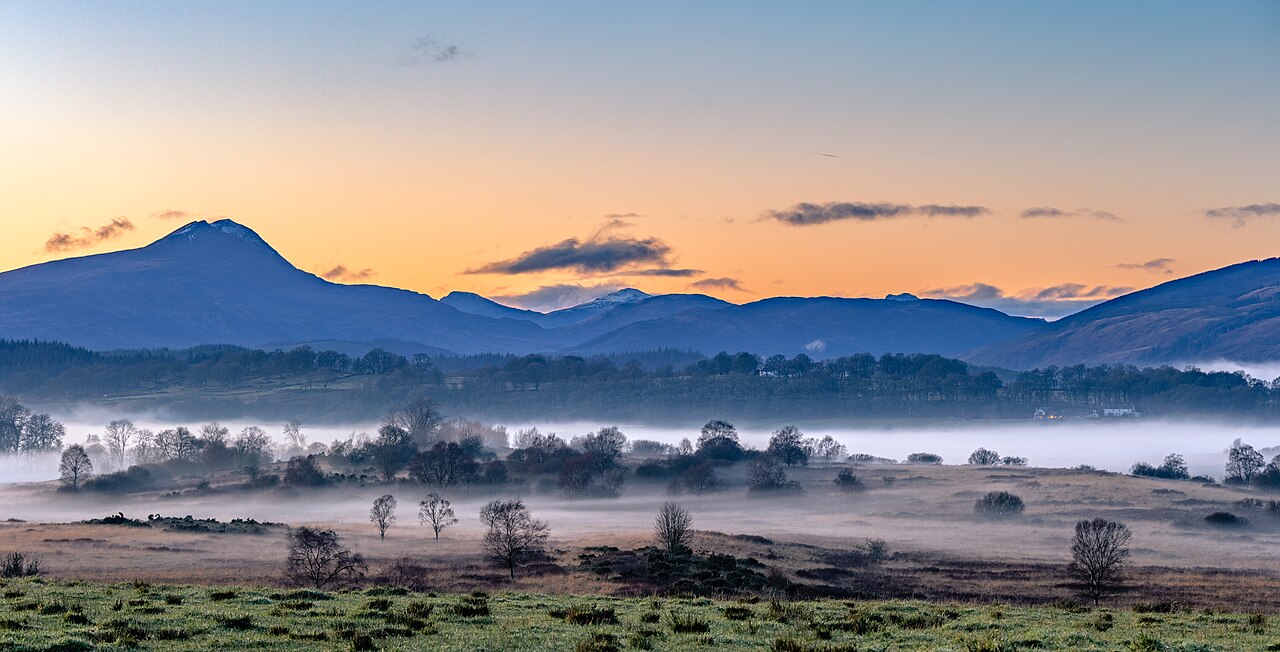 A wide landscape view at sunset showing mist over fields with dark mountain peaks in the distance.