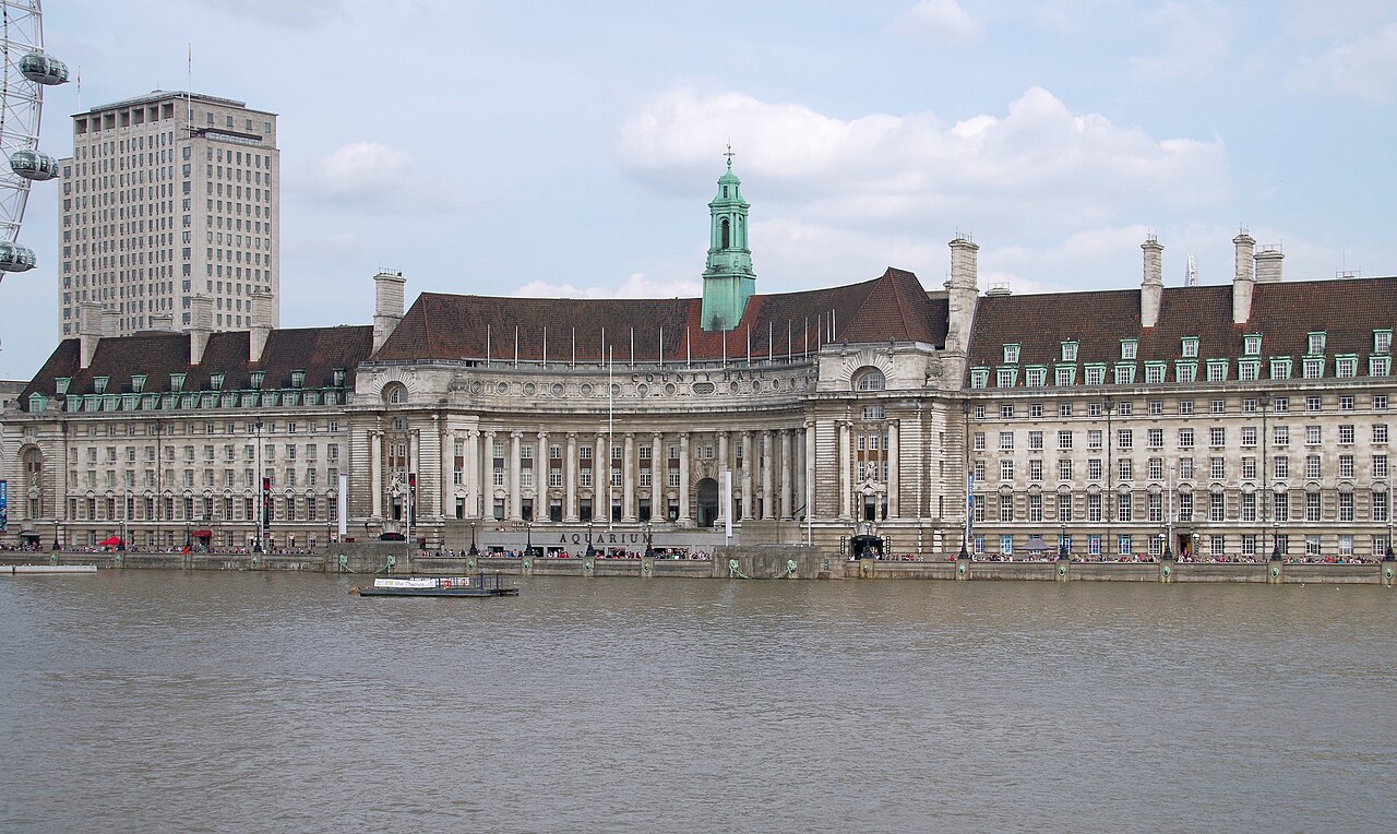 A long curved stone building with a green spire on its roof, fronting a wide grey river.