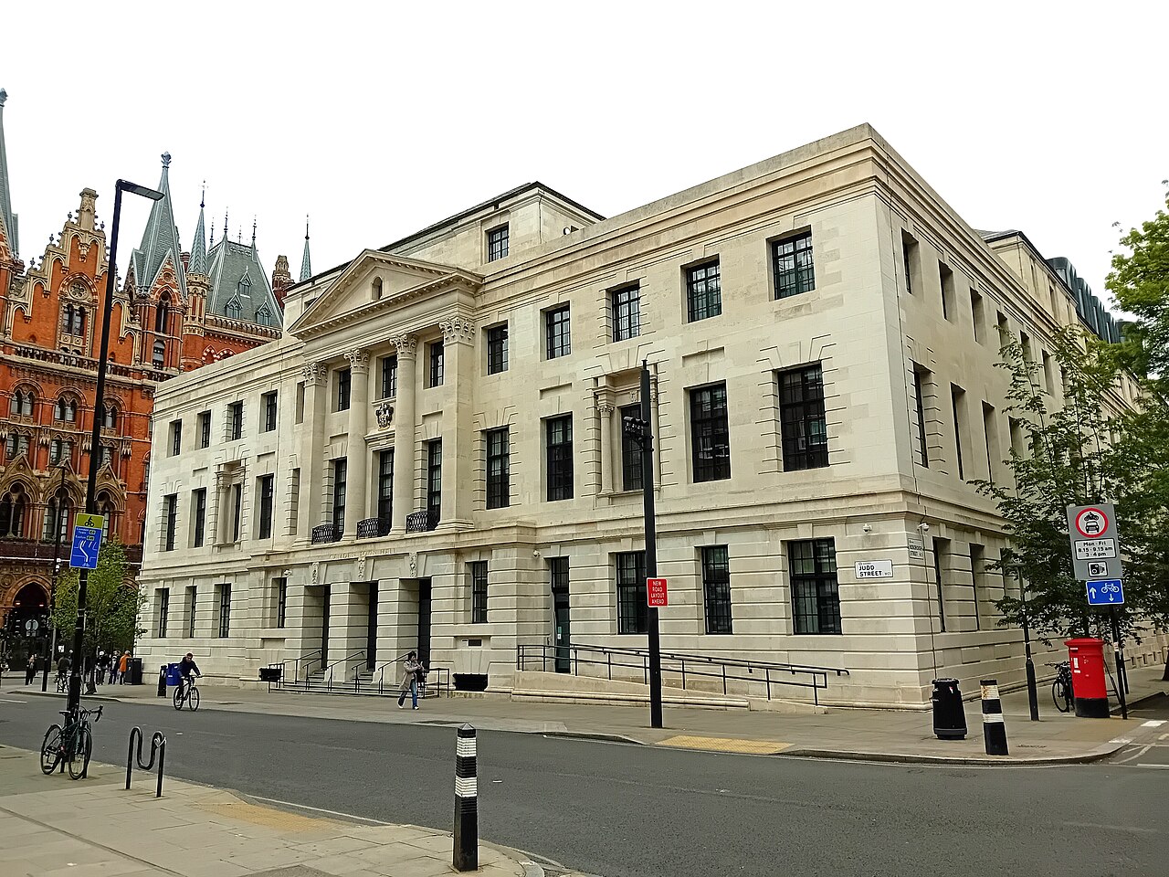 A pale stone civic building with tall windows on a street corner, a red-brick Gothic tower behind it.