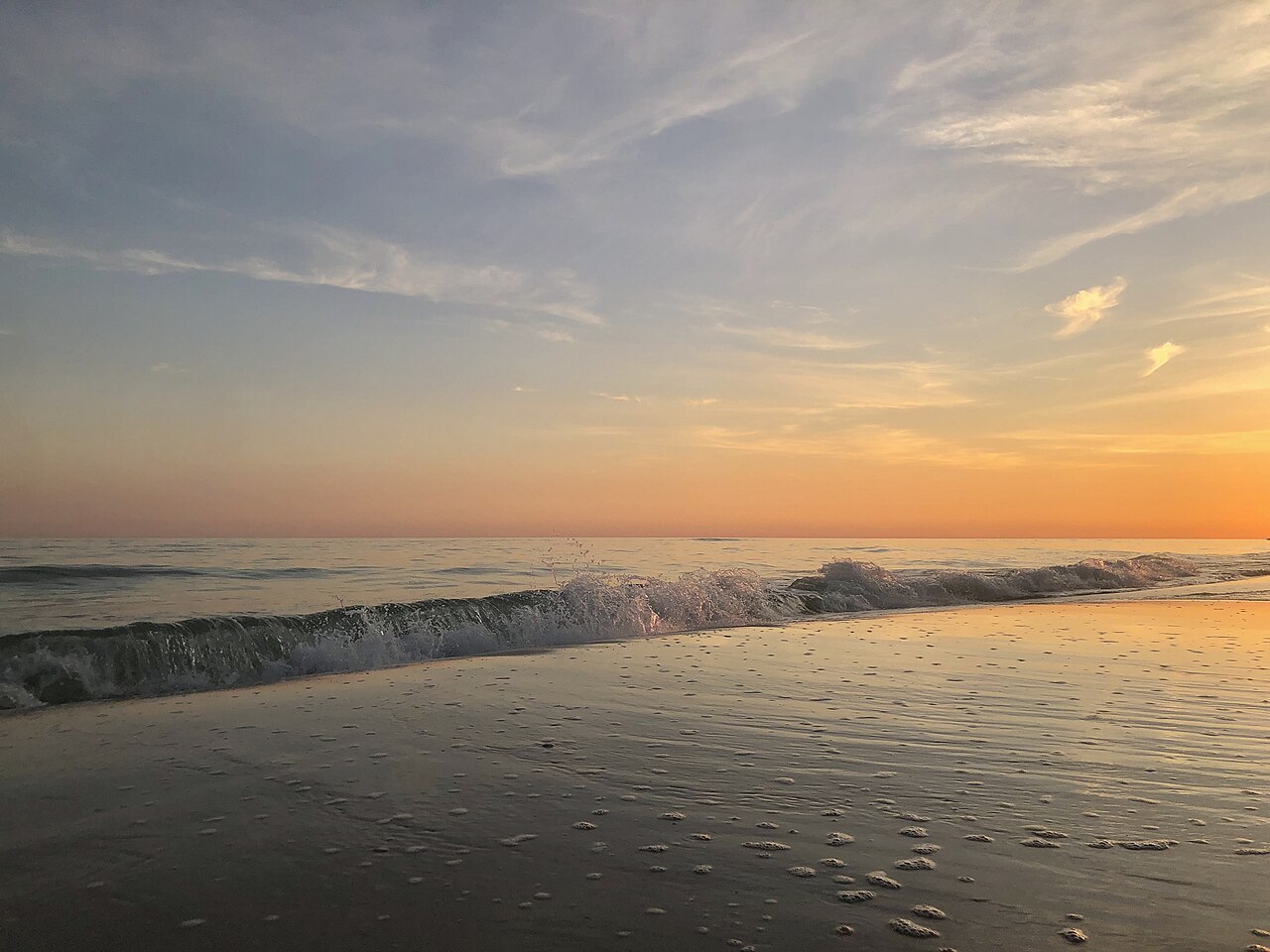 A low wave curling onto wet sand that reflects an orange and grey evening sky.