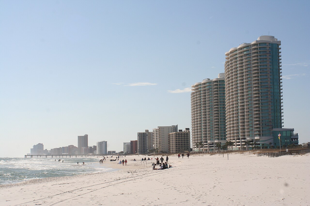A white sand beach curving past a line of tall pale towers under a hazy sky.