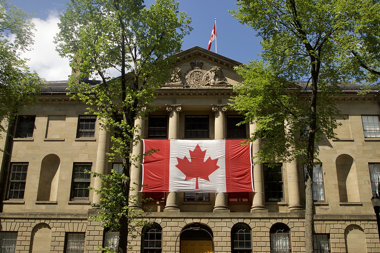 A sandstone building with columns and a pediment, a large Canadian flag hung across its front, framed by green trees.