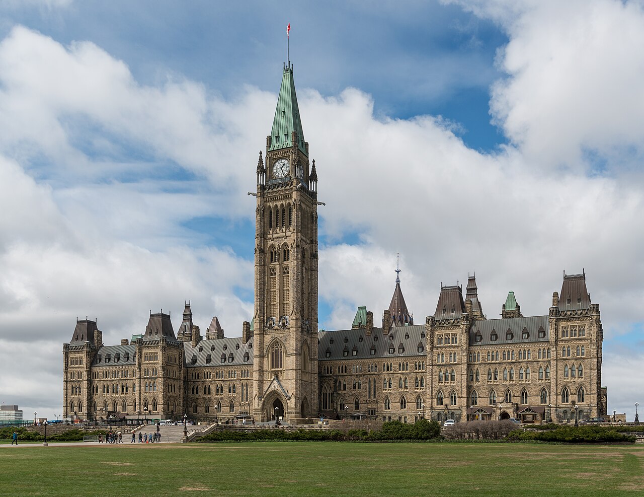 A long Gothic Revival stone building with a tall central clock tower under a sky of broken cloud.