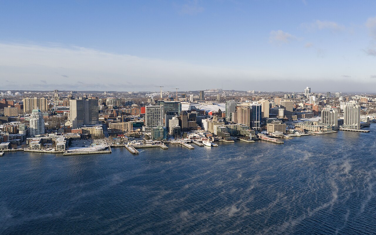 An aerial view of office and apartment towers packed along a waterfront, with dark blue water in the foreground.