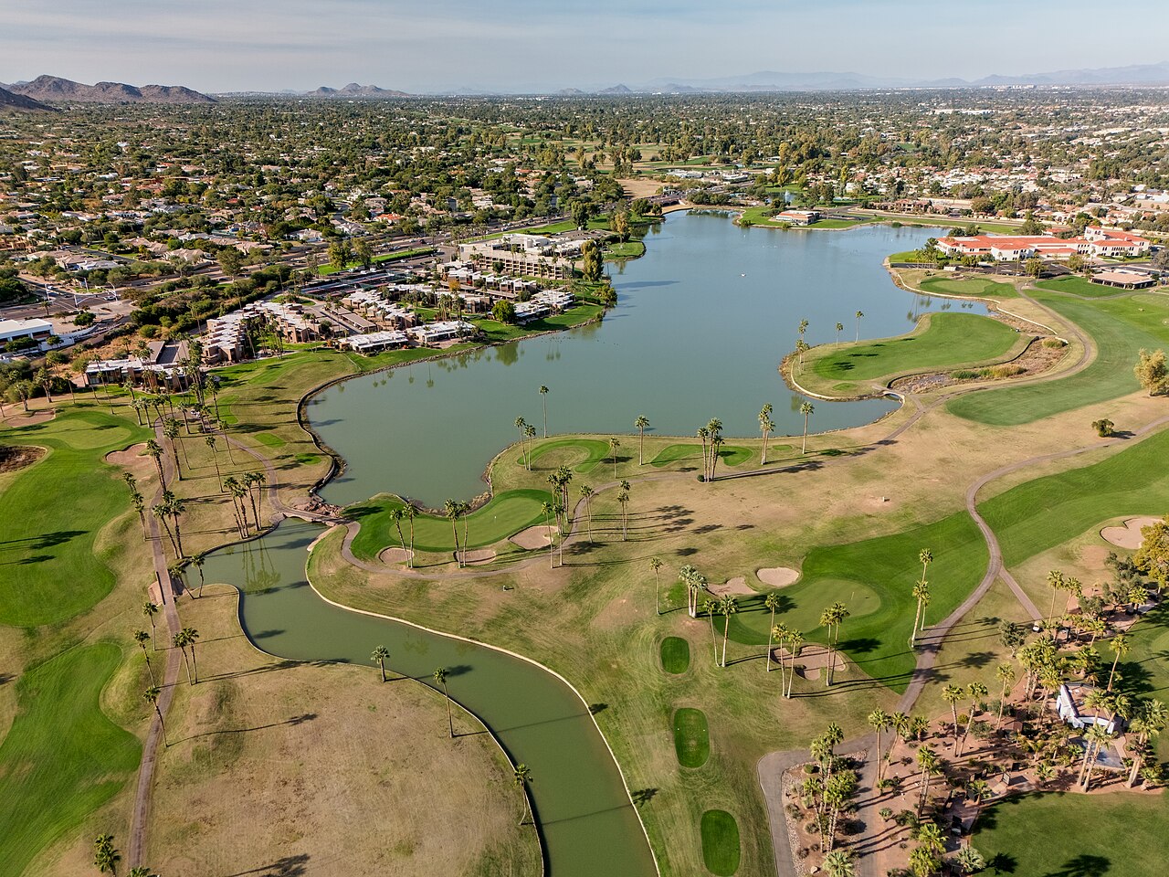 An aerial view of a winding lake and green fairways surrounded by low houses and desert-toned neighbourhoods.