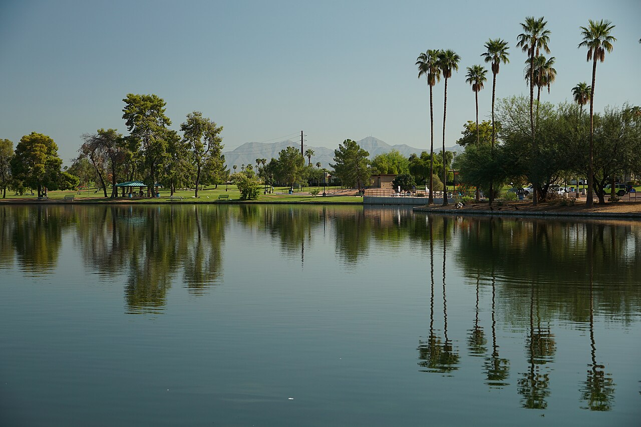A still green lake reflecting a row of tall palm trees, with low mountains in the haze behind.