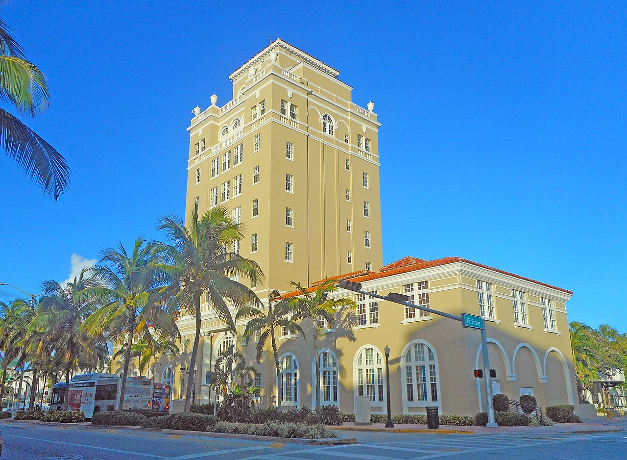 A pale yellow tower of several storeys rising above an arcaded building, with palm trees in front and a deep blue sky.
