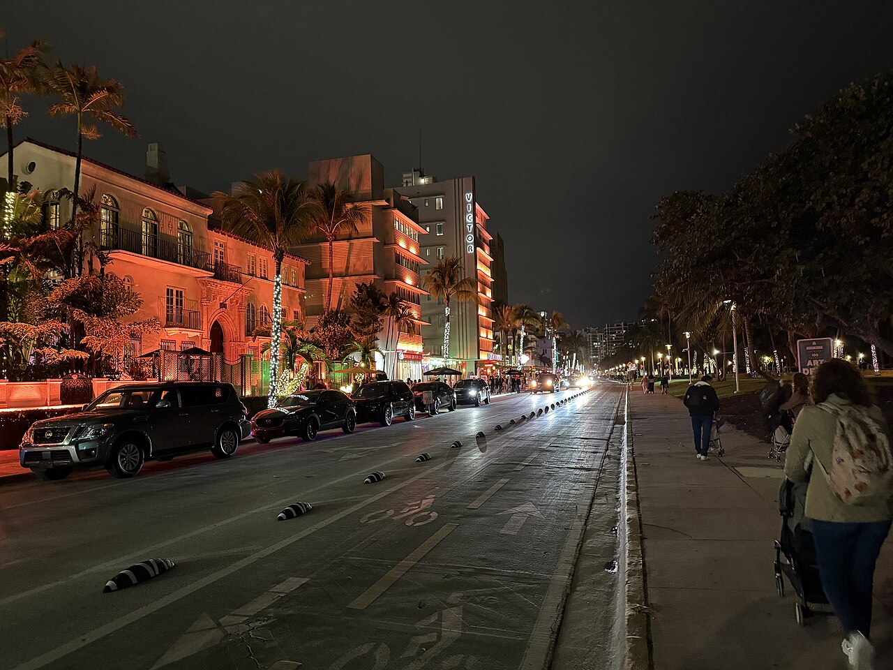 A dark street with parked cars and low buildings lit in orange on the left, and a path beside trees on the right.