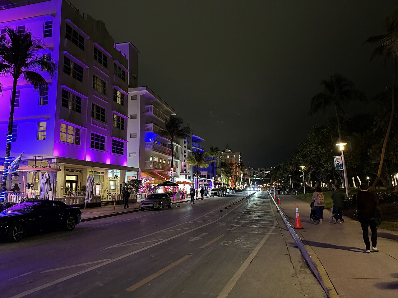 A street at night lined on one side with low hotels lit in purple and blue, palm trees and a few people walking.