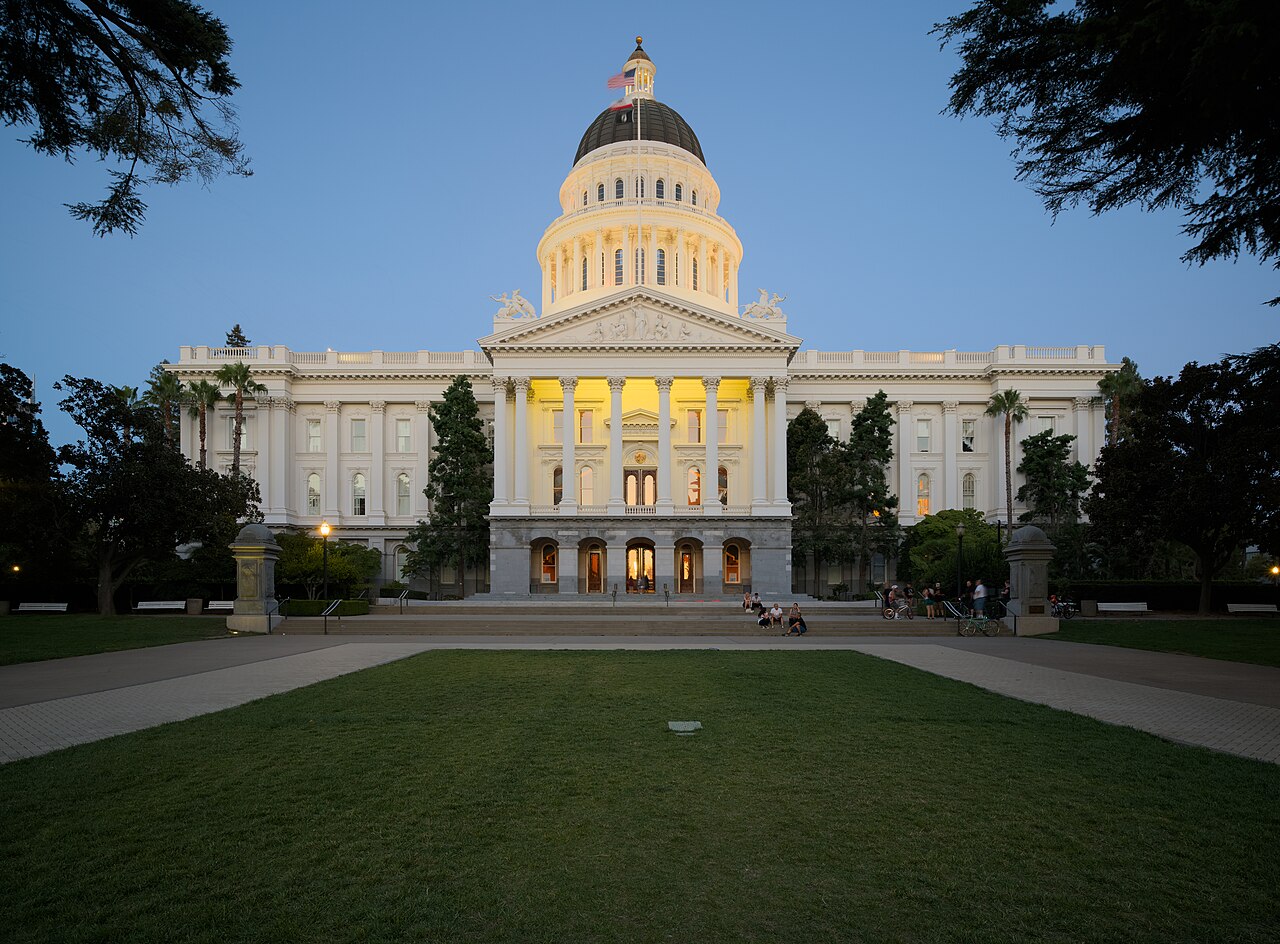 A white domed capitol building with lit windows at the end of a broad lawn, under a deep blue evening sky.
