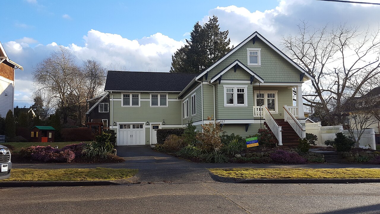 A green two-storey wooden house with white trim, a garage at street level and planted beds along the driveway.