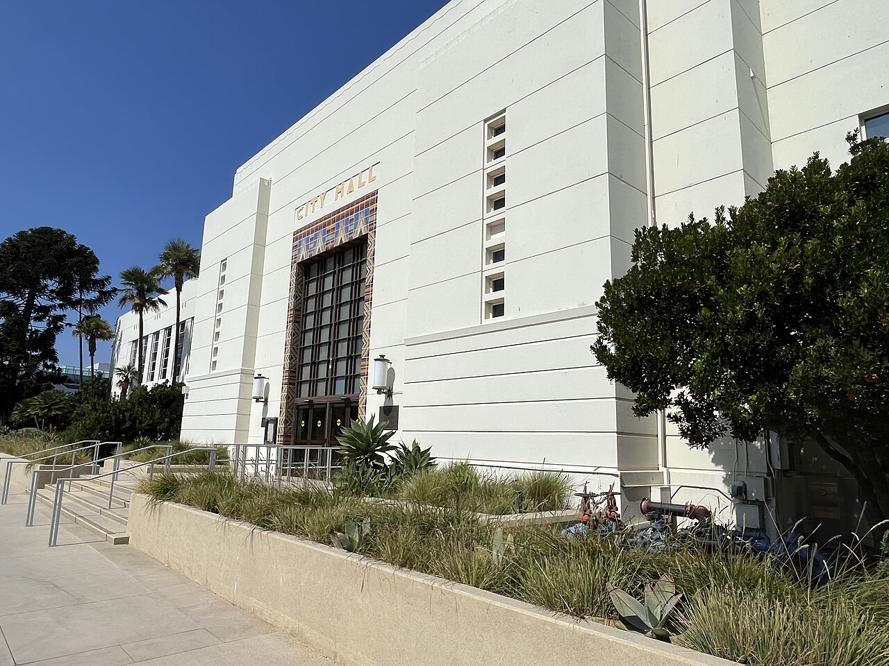 A white Art Deco civic building with a tall recessed entrance, seen at an angle past shrubs and a row of palms.