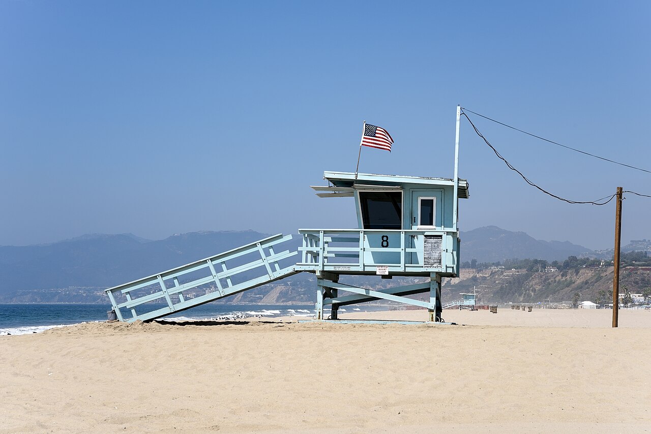 A pale blue lifeguard tower with a ramp and an American flag on a wide empty beach, hills faint across the bay.